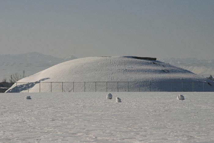 稲荷森古墳の北側の赤湯小学校から稲荷森古墳を見ると雪に覆われ巨大なお供え餅のように見え　その大きさを実感することができます　この稲荷森古墳の南側に南森古墳（仮称）というもっと大きな古墳があったのではと発掘が進められています　古代へのロマンがまた広がります