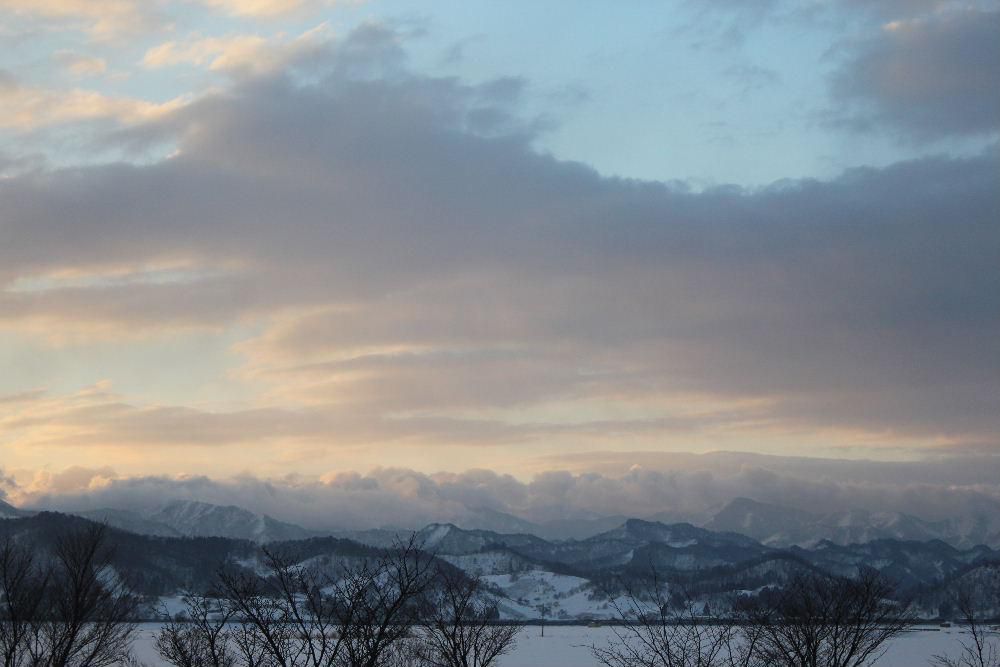 早春の山並み　朝から柔らかな日差しが雪を照らしてくれています　東の空に浮かぶ雲が少し赤みを帯び遠くの山並みを照らしています真っ白だった山が雪と木々がまだらな山を色づけて春らしい感じの山になってきました　少～し春の兆しが感じられる朝のひと時です